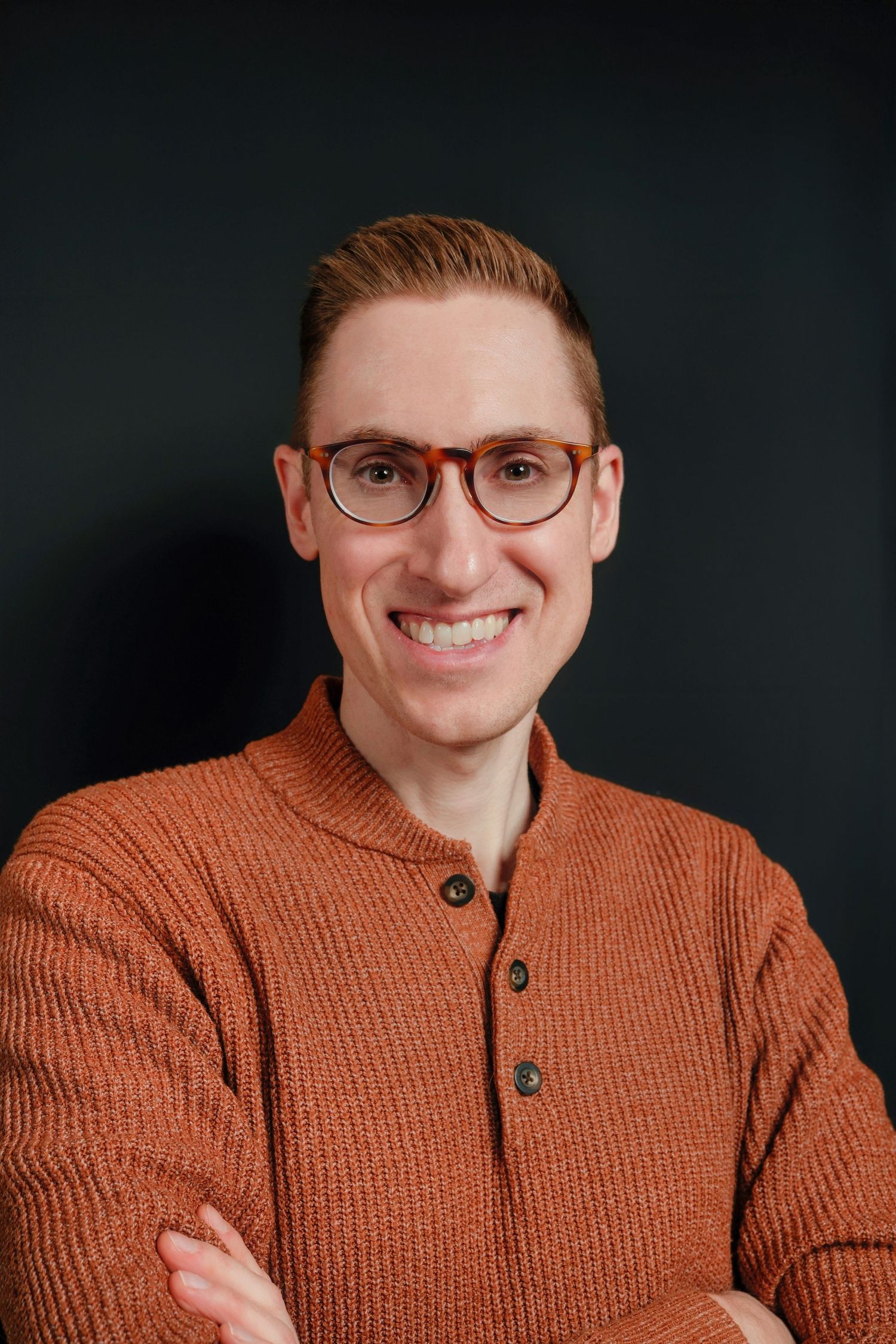 Portrait of a person in knit sweater and glasses smiling against dark background.