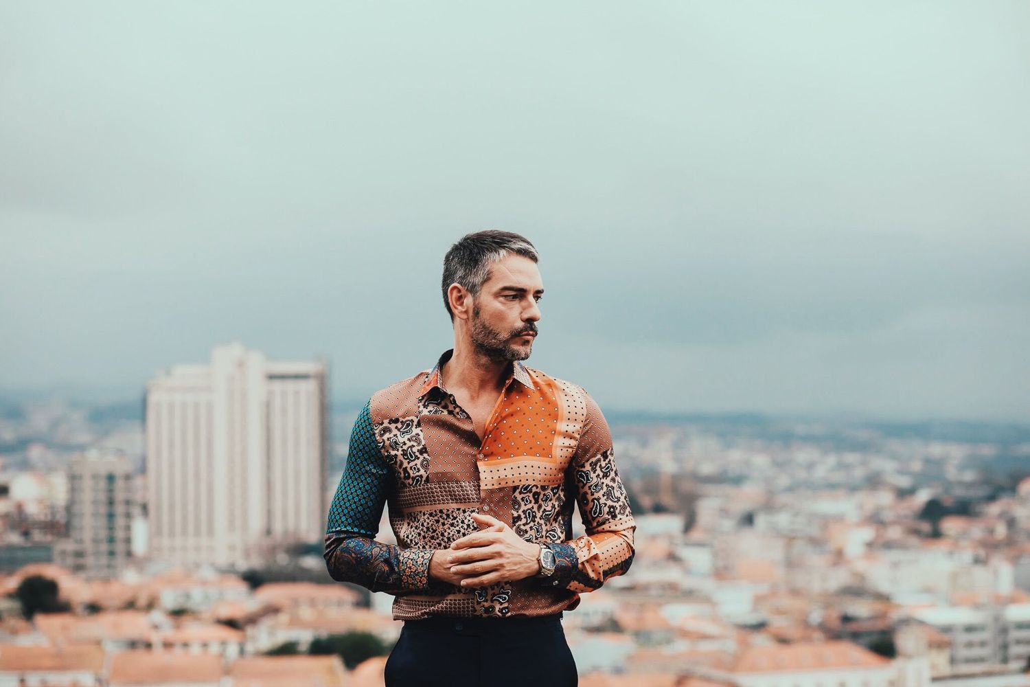 Person with tattoos wearing orange shirt posing against cityscape backdrop on hilltop.