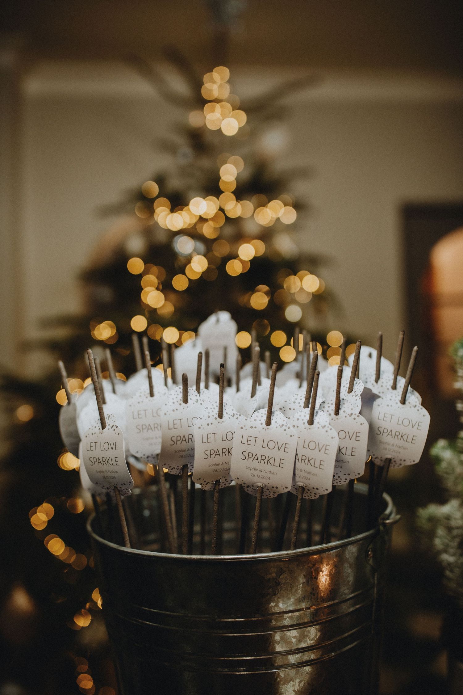 Wedding sparklers with 'Let Love Sparkle' tags displayed in a metal bucket with Christmas tree lights bokeh in background.