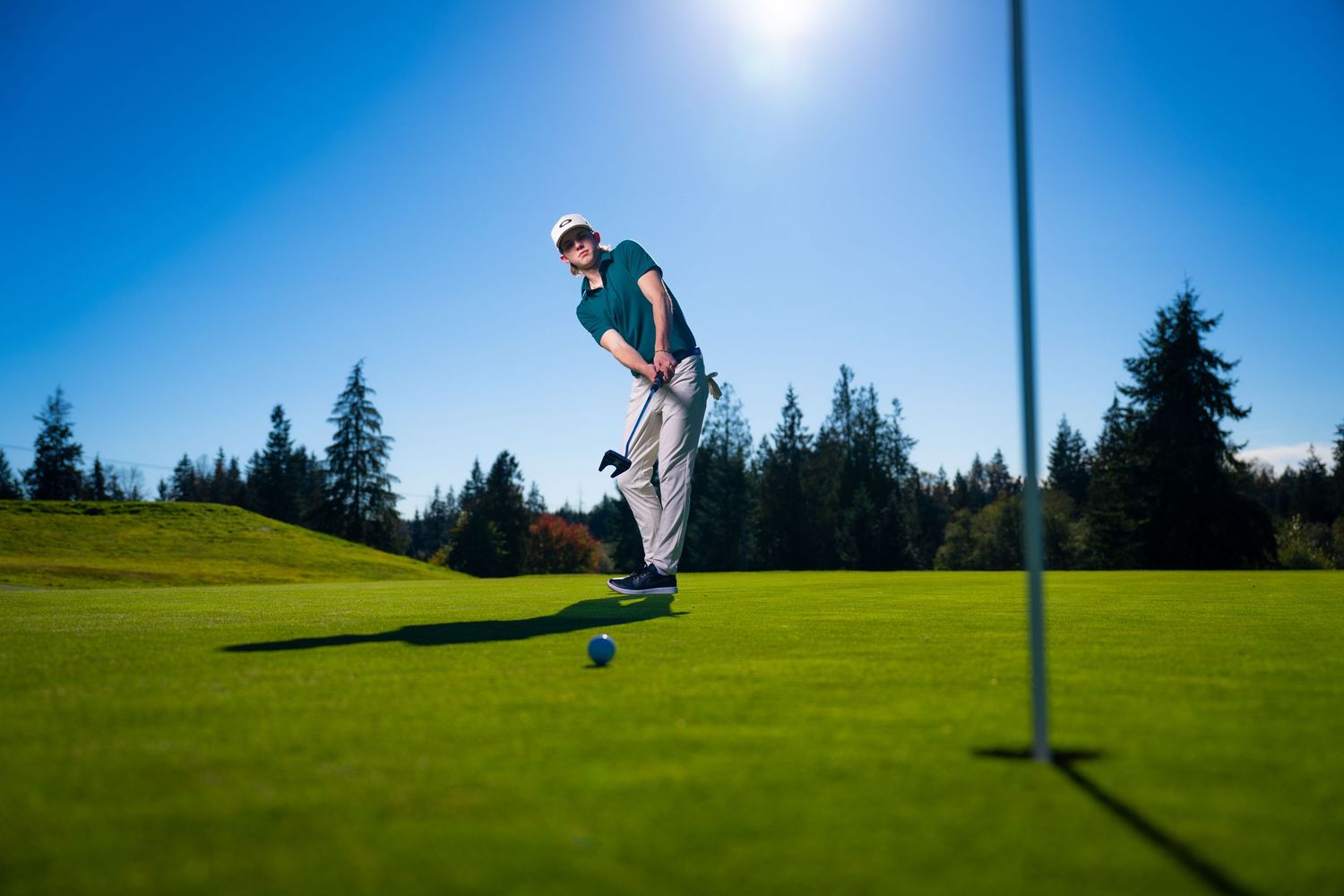 Golfer putting on a vibrant green golf course with evergreen trees and bright blue sky in the background.
