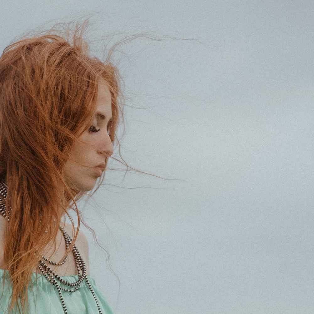 Wind-swept portrait with flowing red hair against minimalist grey background.