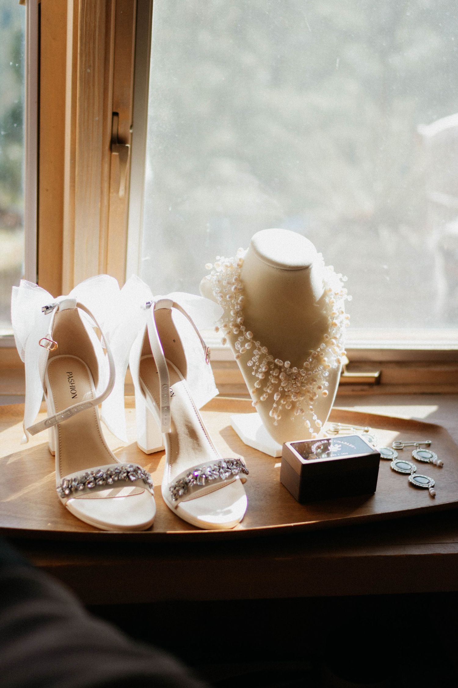 Elegant white bridal accessories including heeled sandals, pearl necklace and jewelry displayed on wooden windowsill. Wedding detail picture