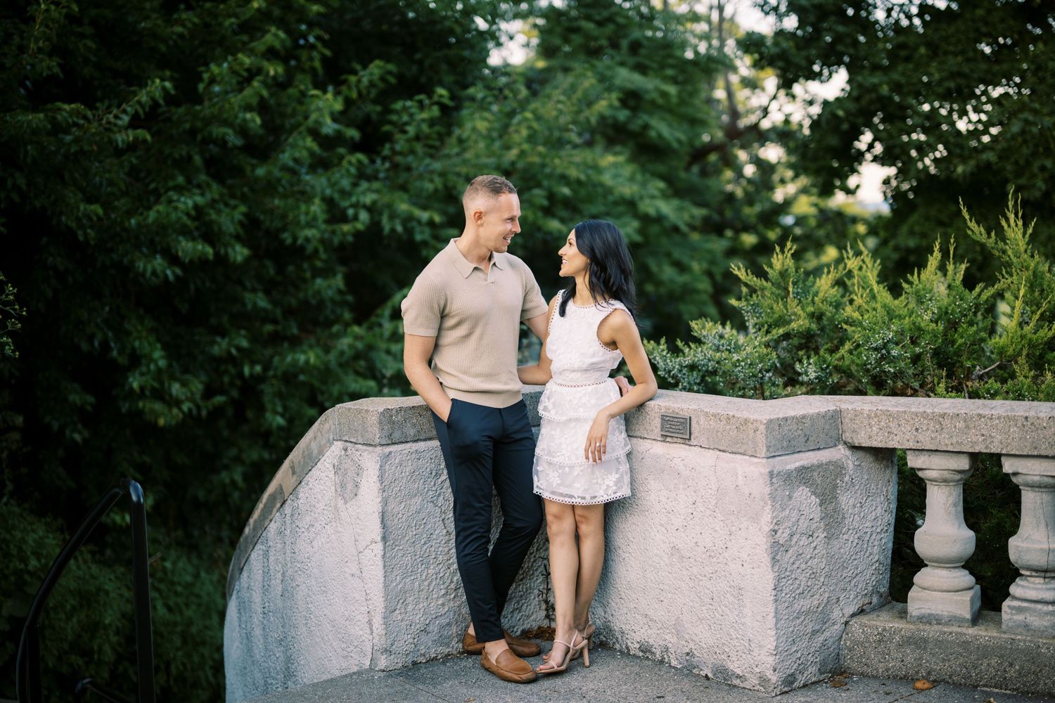 Couple stands together on a stone bridge balcony surrounded by lush green foliage during a romantic outdoor photo session.