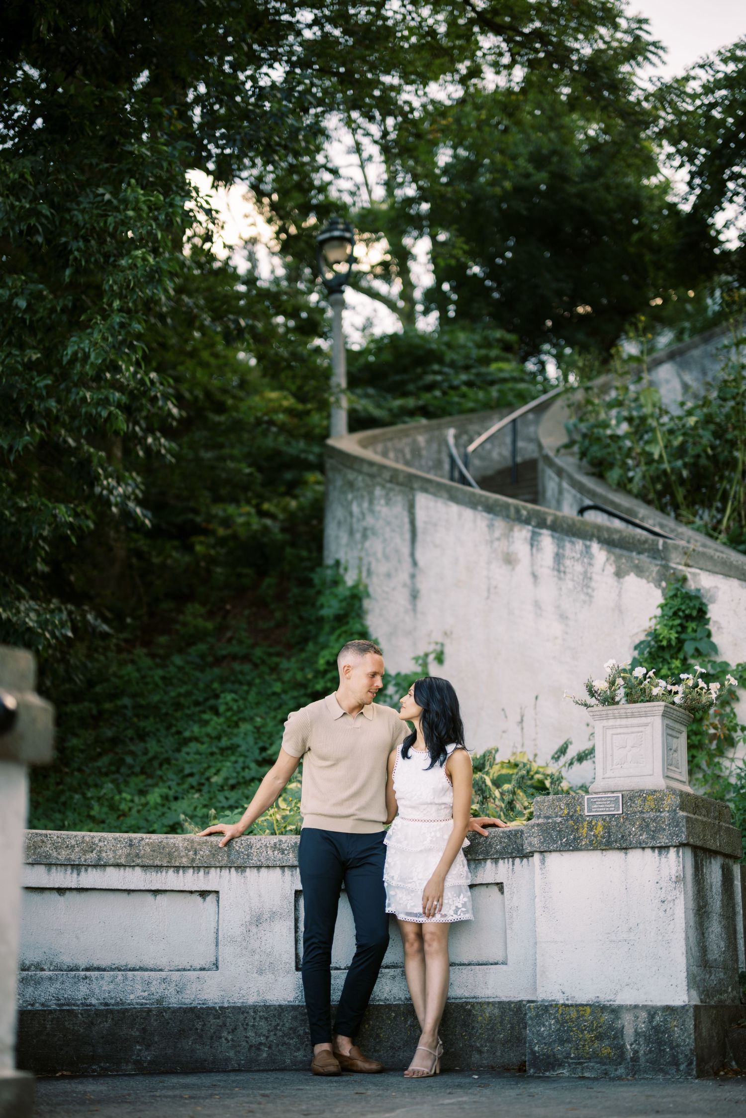 A couple in casual attire stands together on concrete steps surrounded by greenery at dusk.