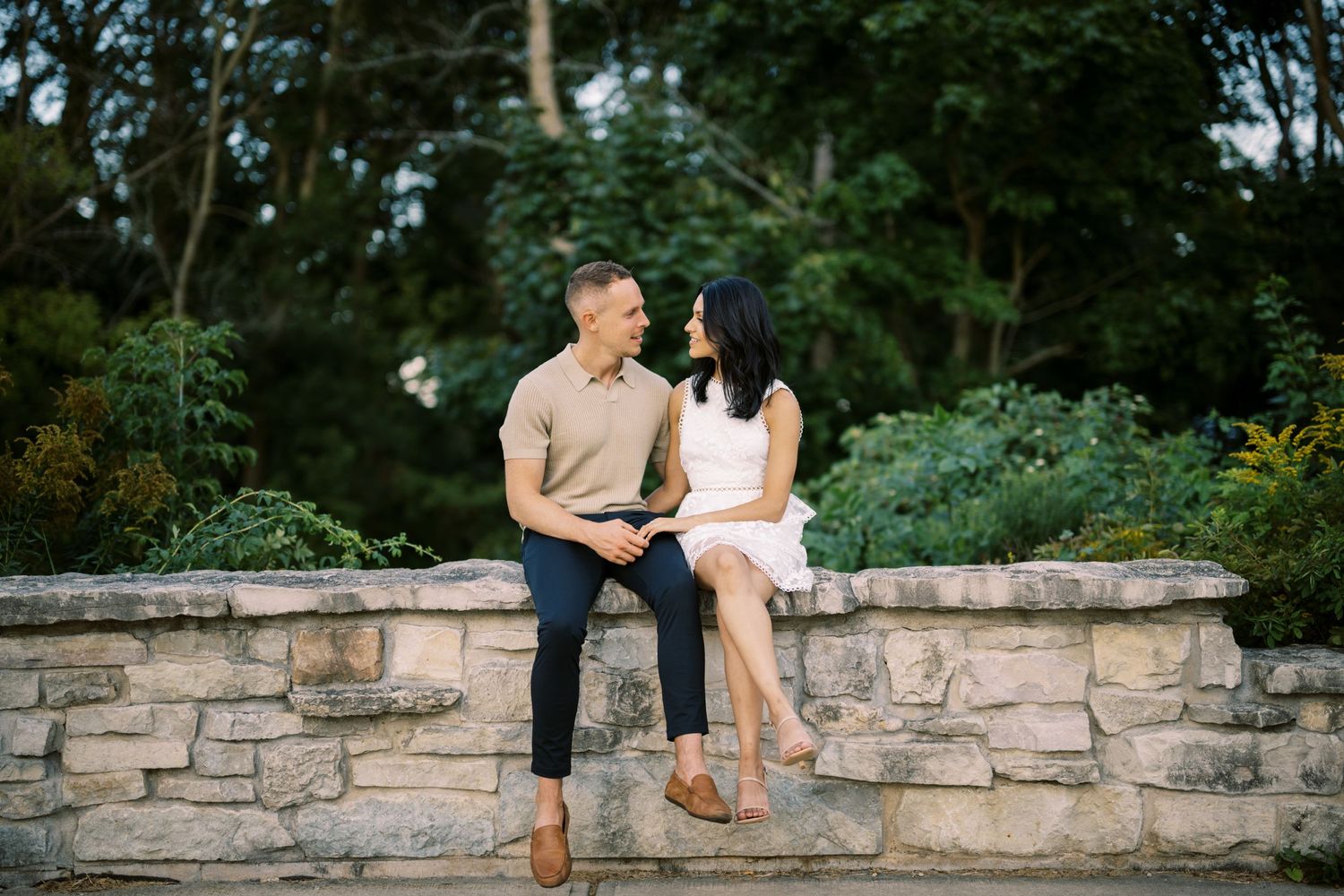 Two people sit together intimately on a stone wall in a garden setting during a romantic photo session.