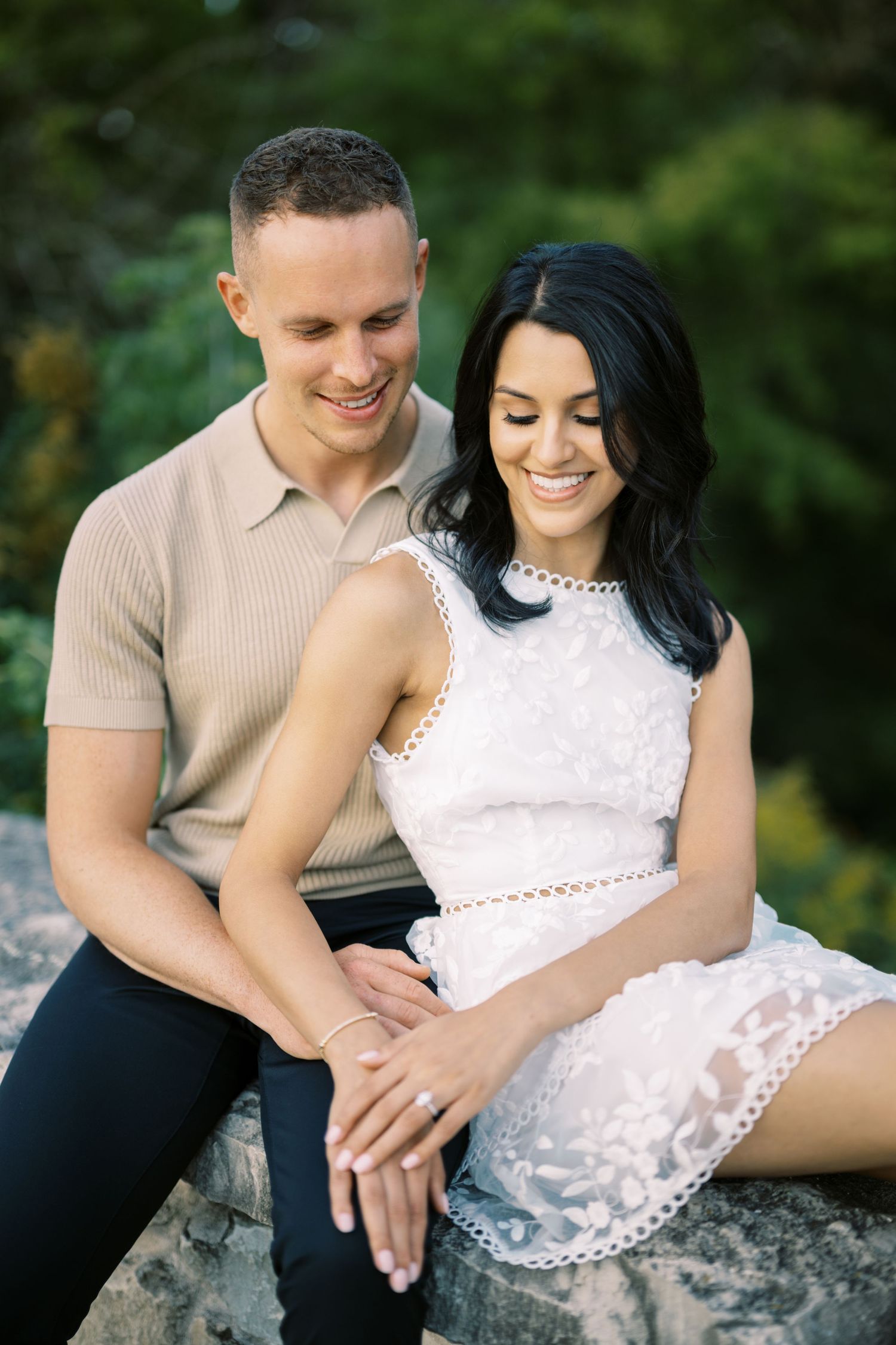 A couple in casual summer attire sits close together on a rock outdoors, sharing an intimate moment in a natural setting.