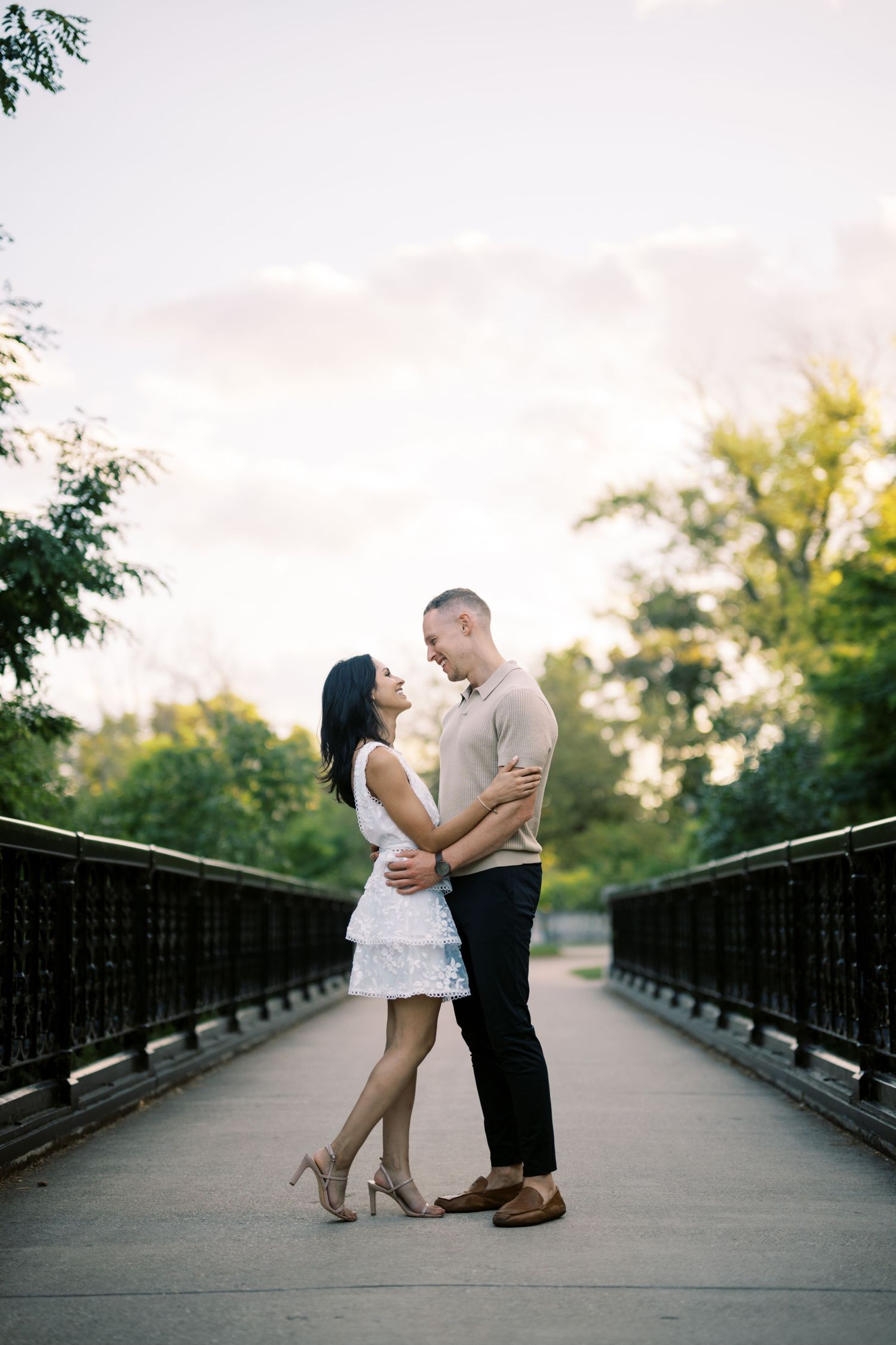 A couple stands embracing on a wooden bridge during sunset with lush greenery in the background.