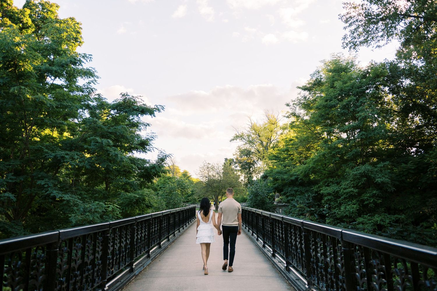 A couple walks hand in hand across a scenic bridge surrounded by lush green trees at sunset.