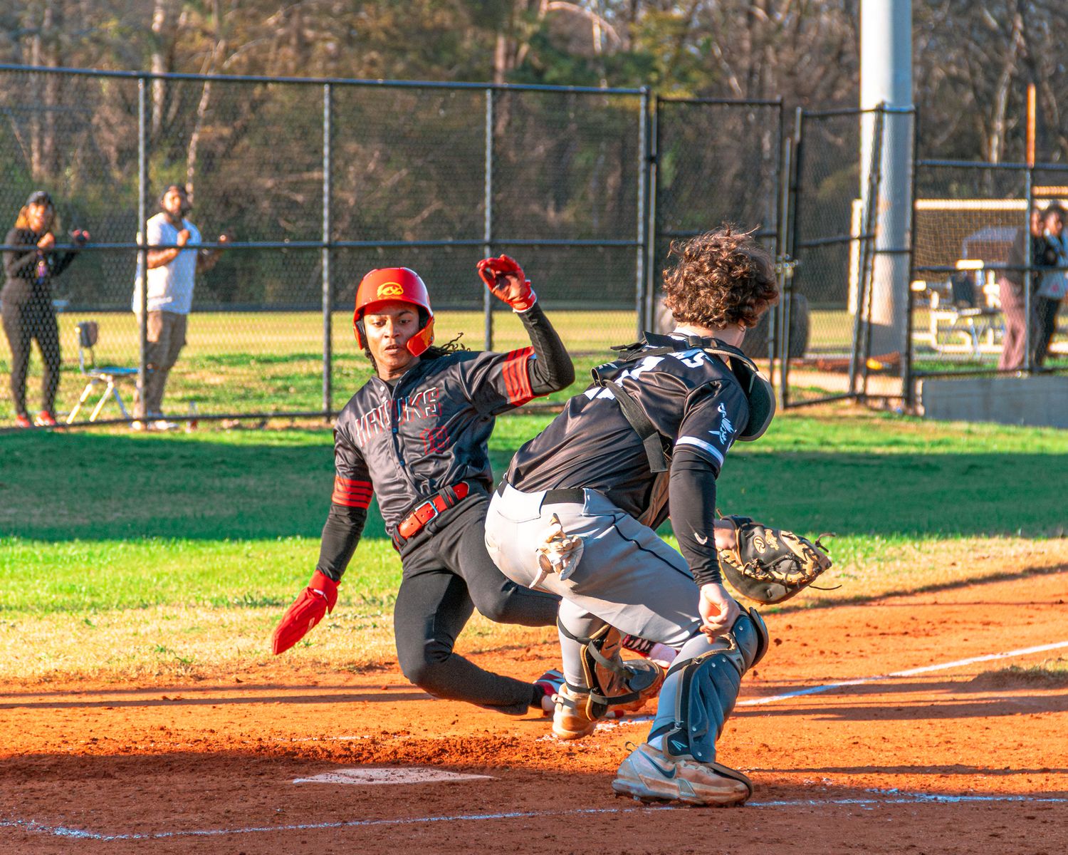 Dramatic baseball play at home plate as runner slides past catcher during game on sunny afternoon.