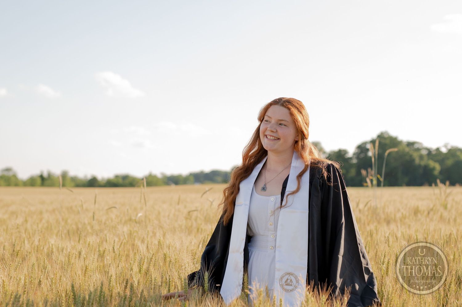Graduate in cap and gown poses in golden wheat field during sunset.