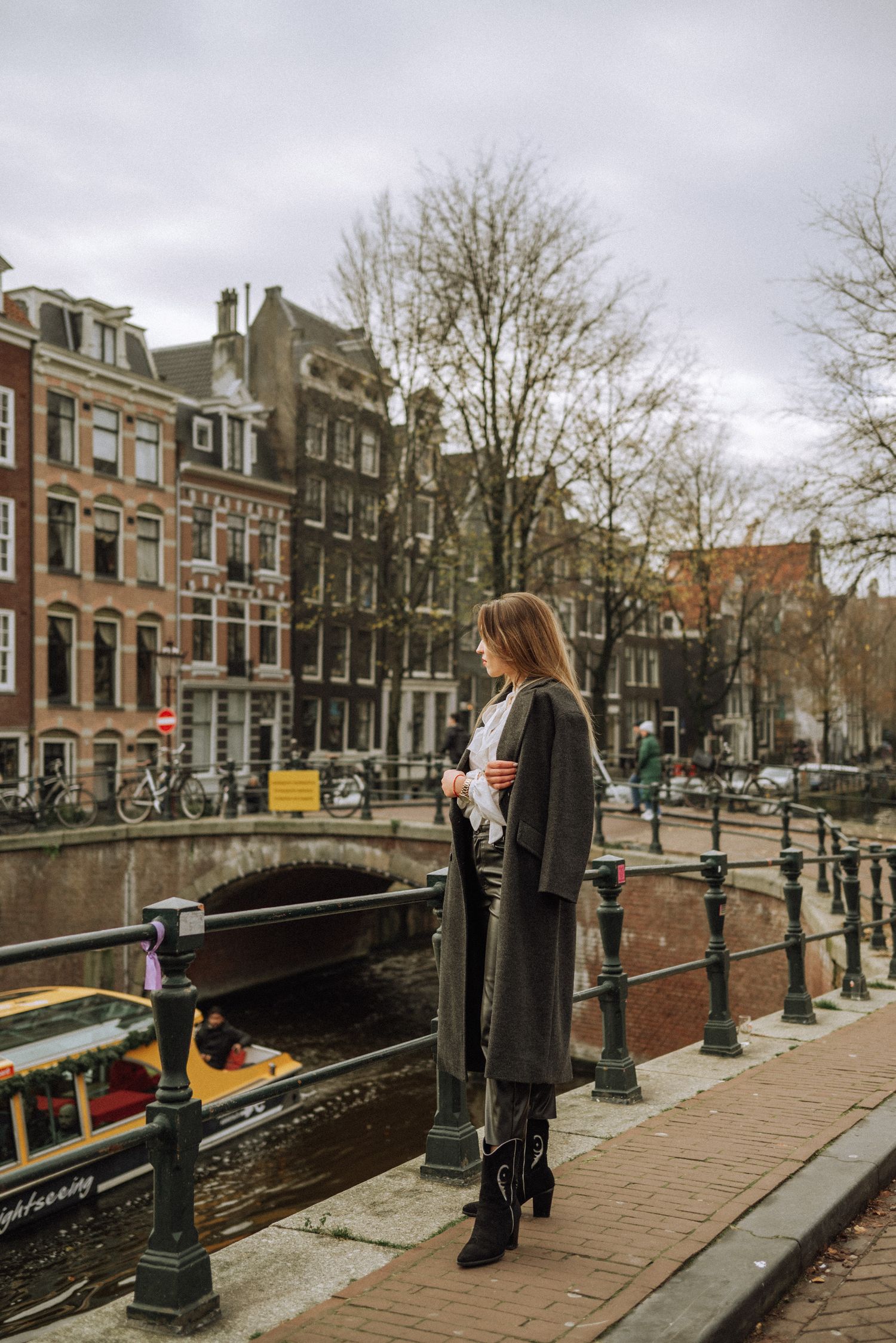 A woman during a solo photoshoot in Amsterdam standing on a bridge, watching a boat pass through the canal.