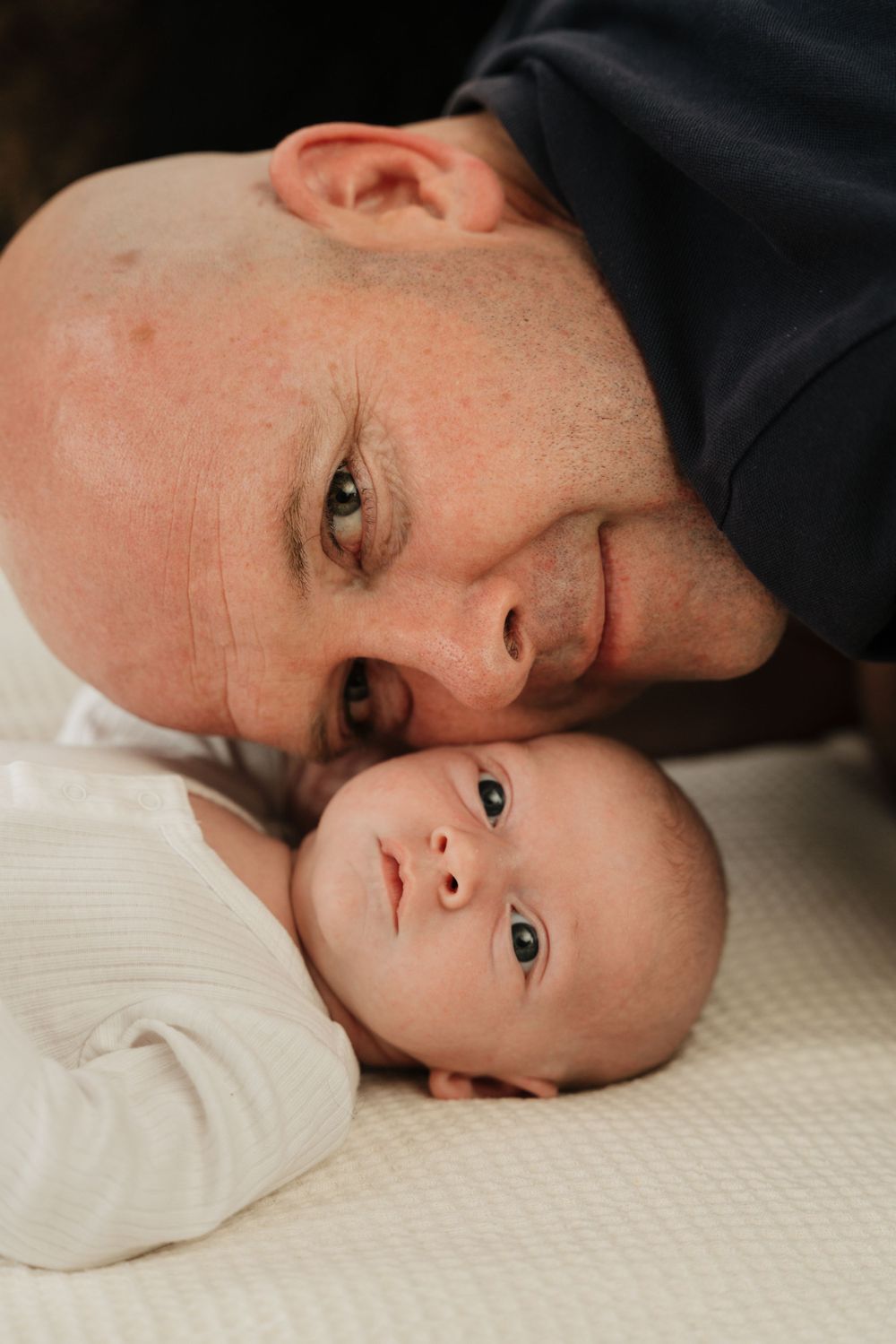 Close-up portrait of parent and baby laying side by side on white sheets.