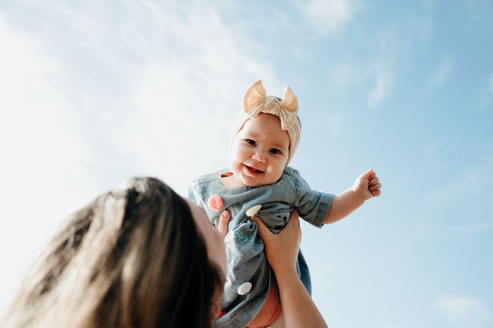 Baby wearing blue outfit and bow held up against bright blue sky.