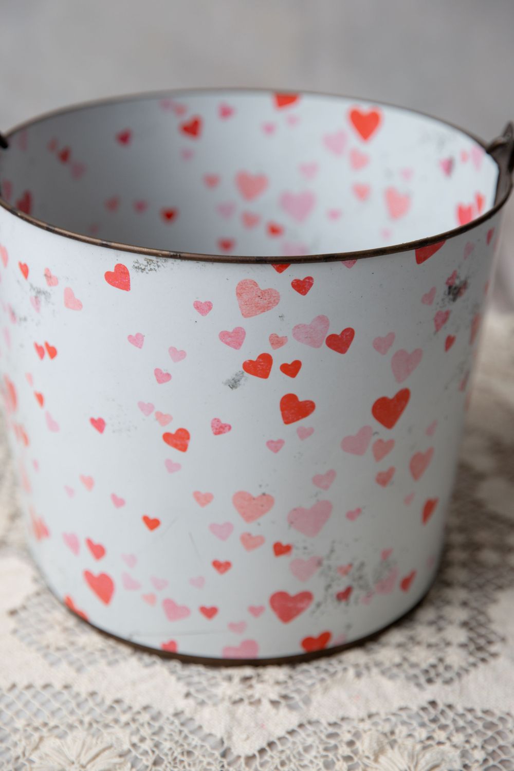 White enamel bucket with red and pink heart pattern, vintage-style metal pail with wire handle on lace surface.