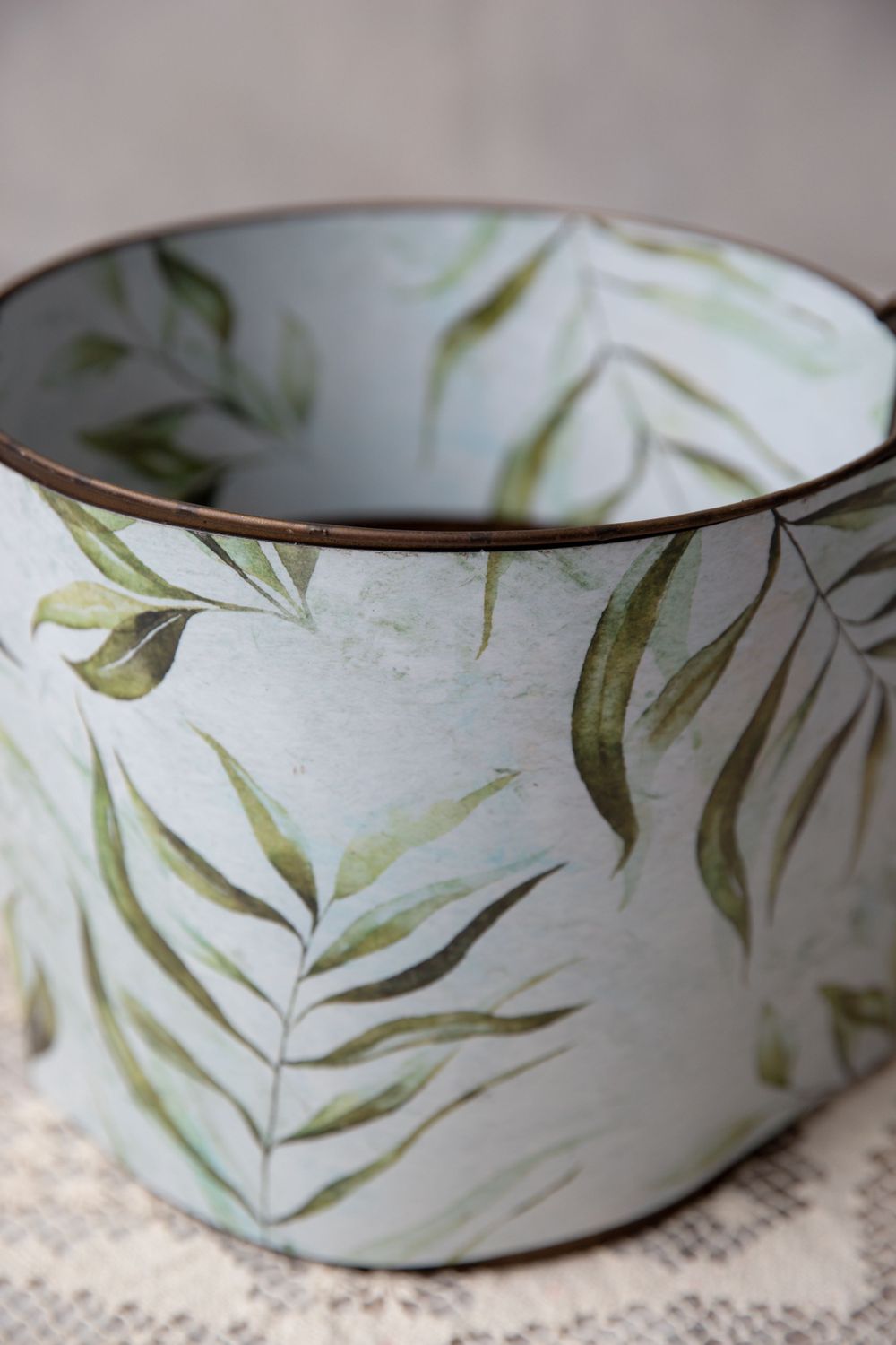 Cylindrical metal container with watercolor tropical leaf pattern on a lace doily surface.
