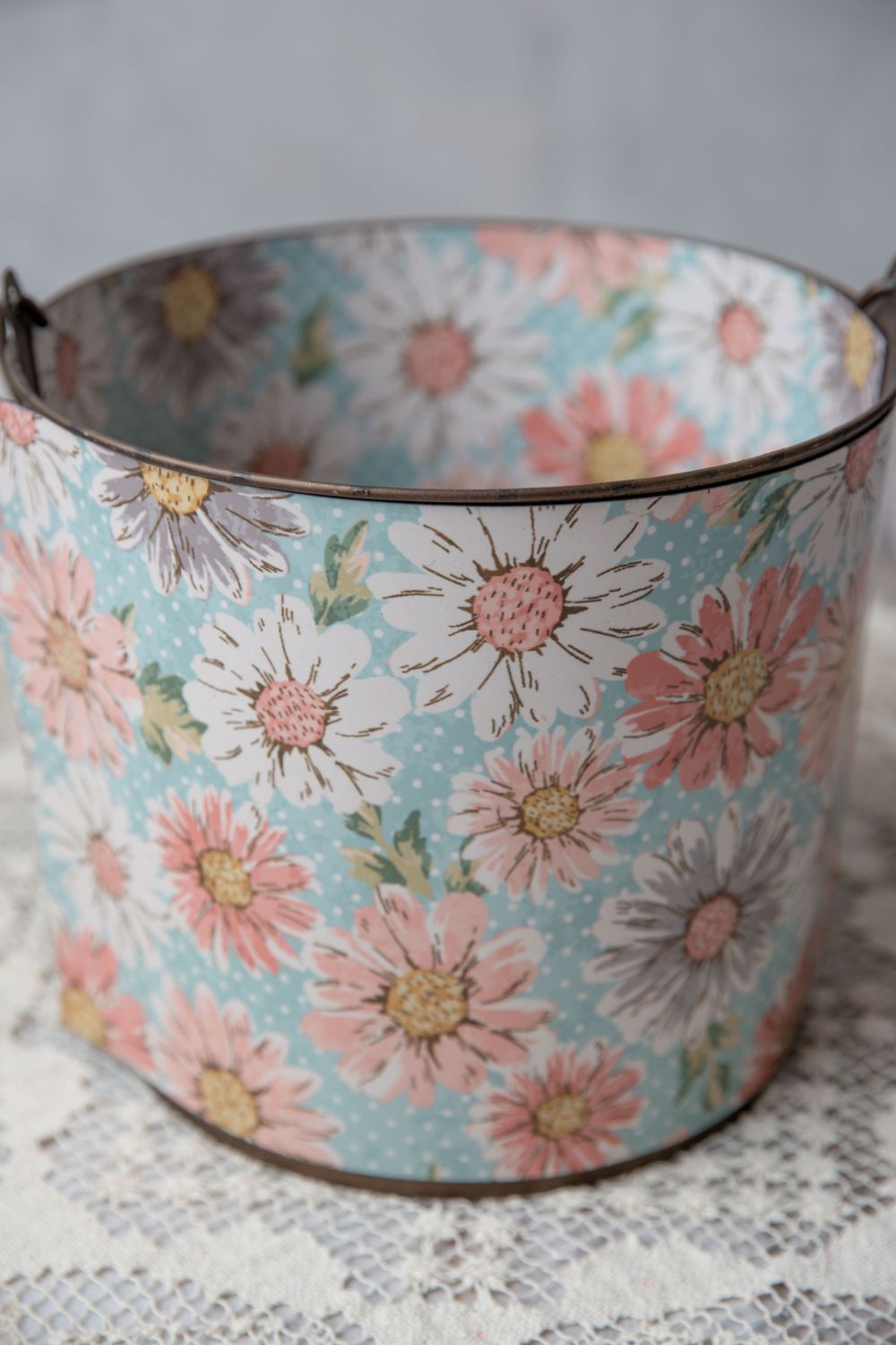 Vintage floral tin bucket with pink and white daisy pattern on light blue polka dot background with metal handle.