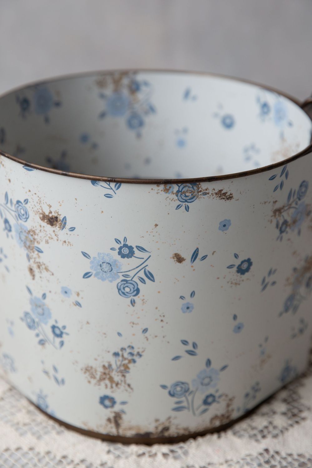 Vintage white tin container with blue floral pattern and rust spots, sitting on lace fabric.