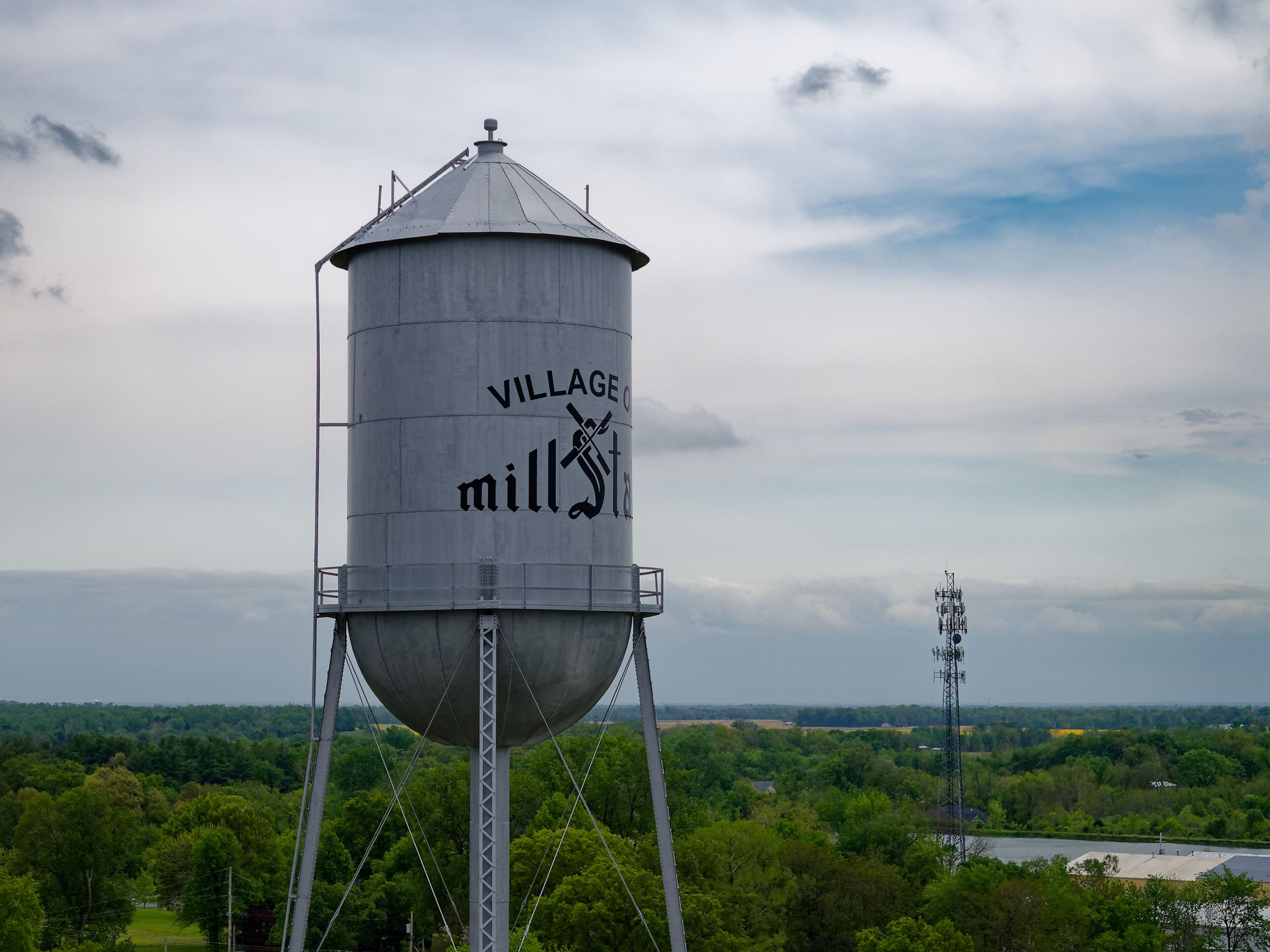 A tall vintage water tower labeled 'Village Mill' stands against a cloudy sky overlooking a green rural landscape.