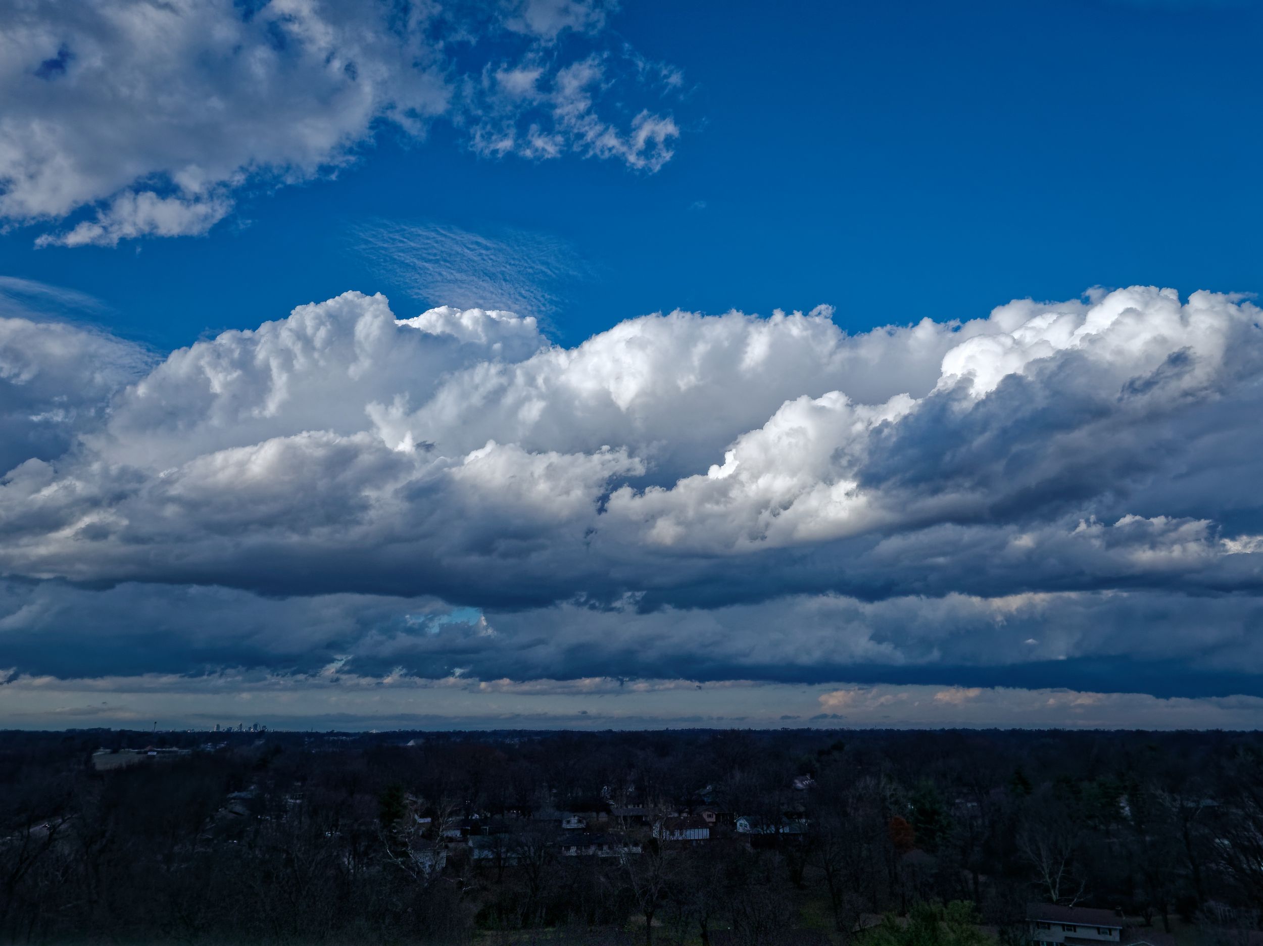 Panoramic view of dramatic white clouds against deep blue sky over a dark forested landscape.