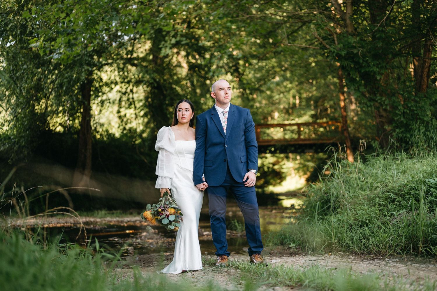 Newlywed couple walking by stream in lush green garden setting.
