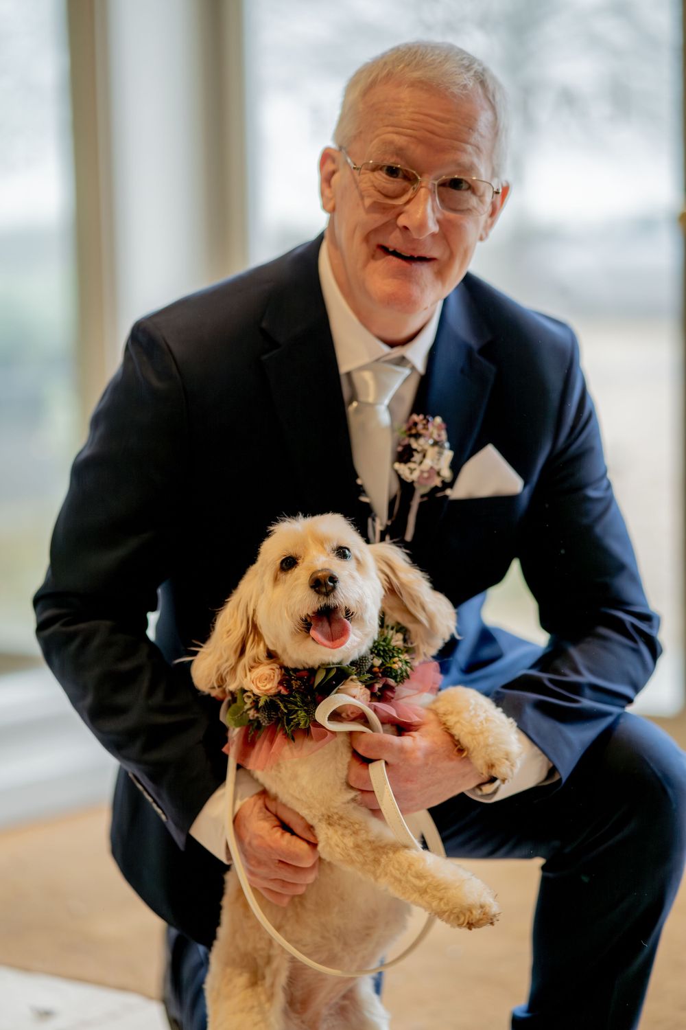 An adorable small dog wearing a floral collar poses with someone in a navy suit at a formal event.