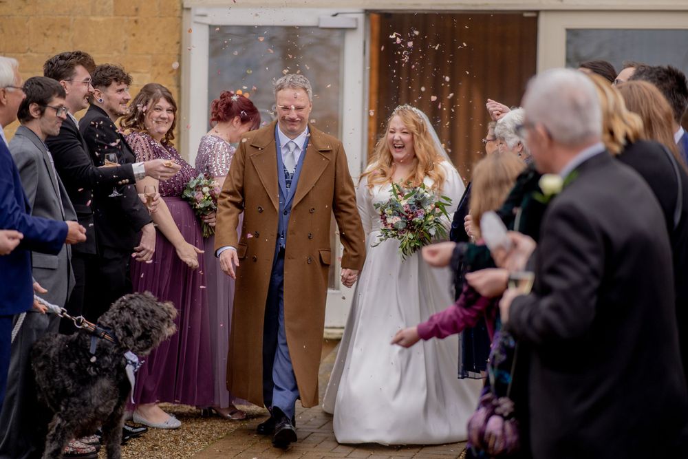 Wedding guests celebrate with confetti as a newly married couple exits church with their dog present at the ceremony.