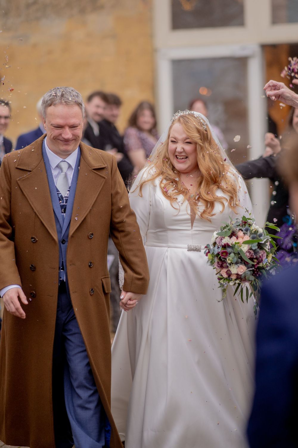 Happy wedding couple walking together outdoors as guests throw confetti, bride in white dress with flowers.