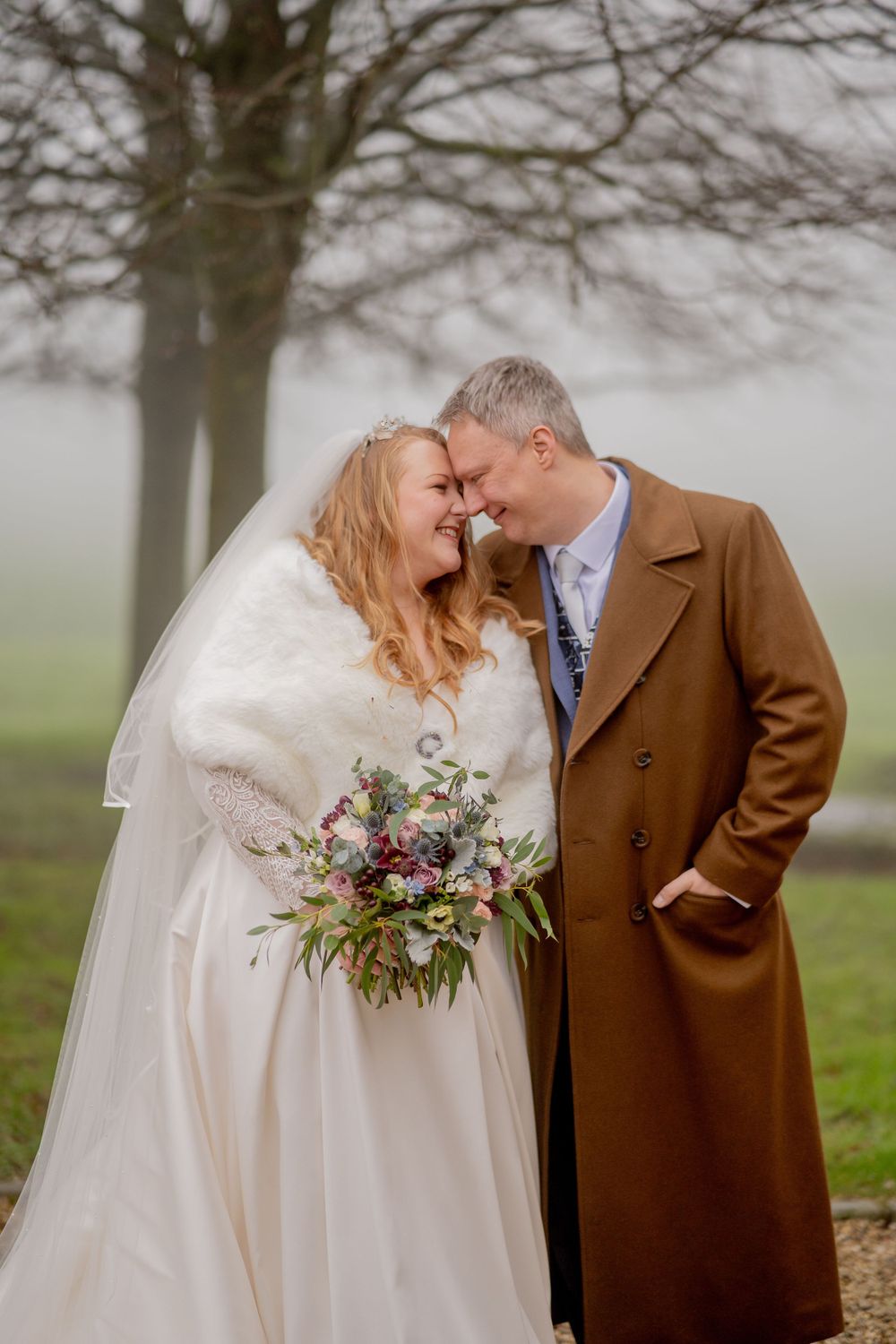 Two people share an intimate winter wedding moment in the foggy countryside wearing formal attire.