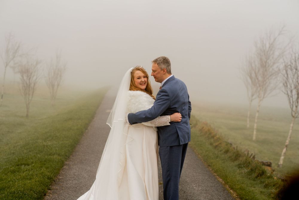 Newlyweds embrace on a misty path surrounded by bare trees on their foggy wedding day.