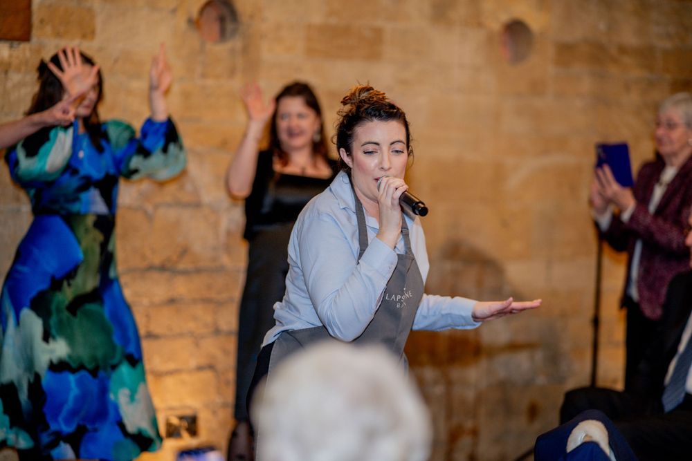 Performer gestures emotively during theatrical performance with stone wall backdrop while others applaud.