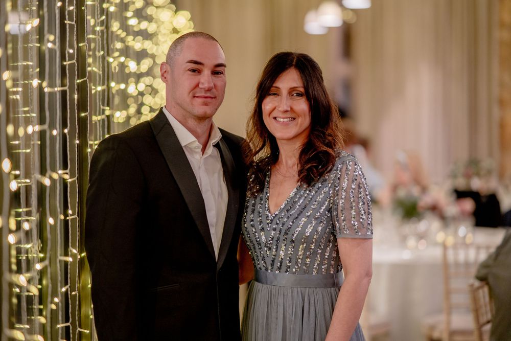 A couple stands together at an evening event with string lights creating a warm ambiance behind them.