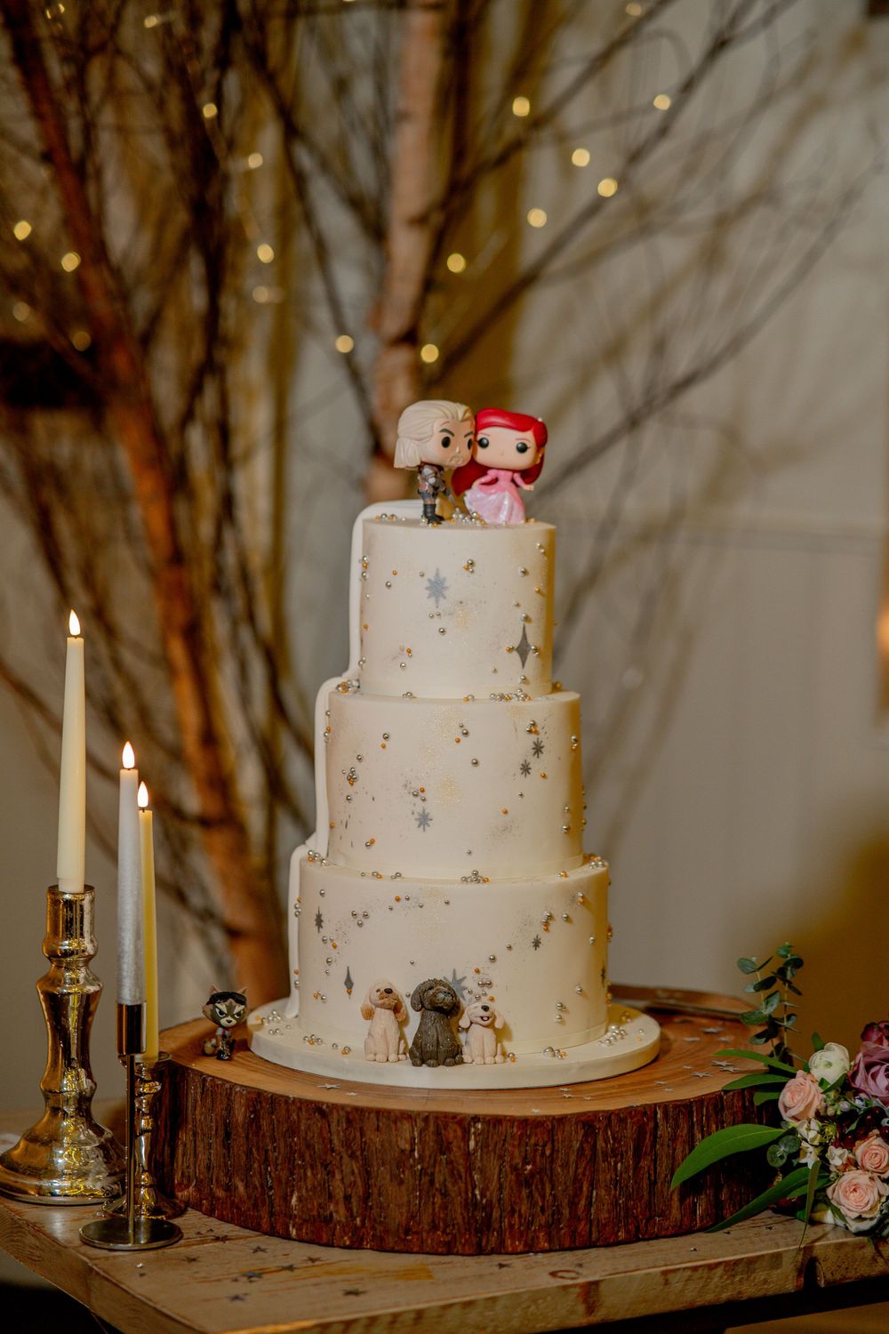A white three-tiered wedding cake topped with pop figurines sits on a rustic wood slice with candles and flowers nearby.