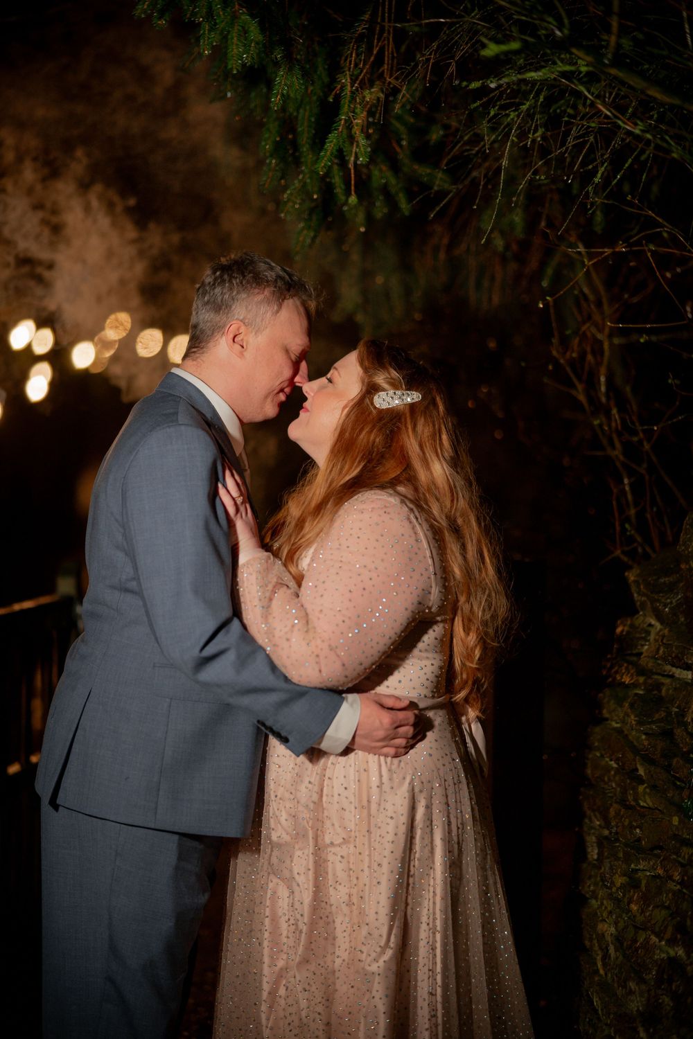 A romantic evening wedding portrait with the couple embracing under twinkling lights and greenery.