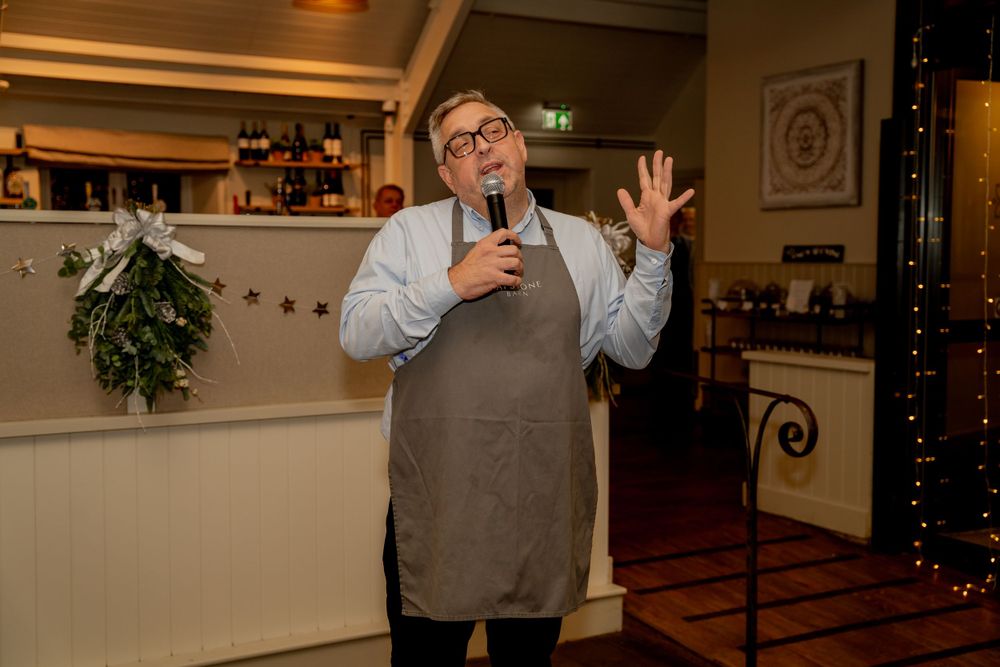 A speaker in an apron gestures animatedly while giving a presentation in a decorated restaurant setting.