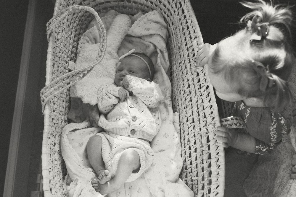 Black and white photograph of a baby lying in a woven bassinet with a cat looking in from the side.