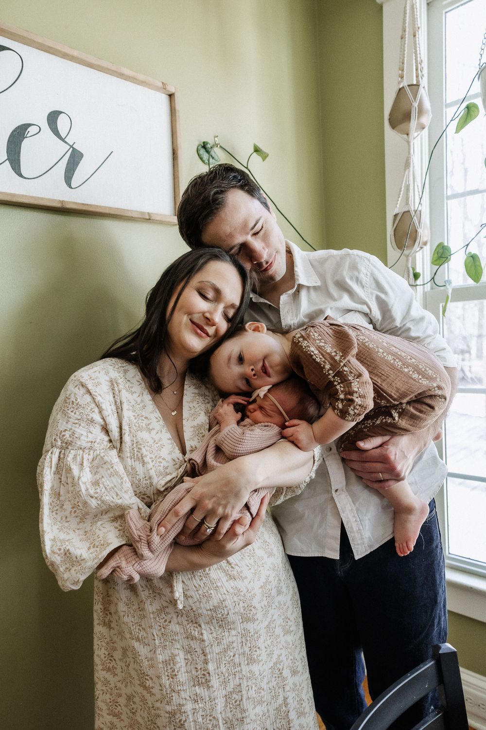 A family shares a tender moment together by a window in a room with sage green walls and natural light.