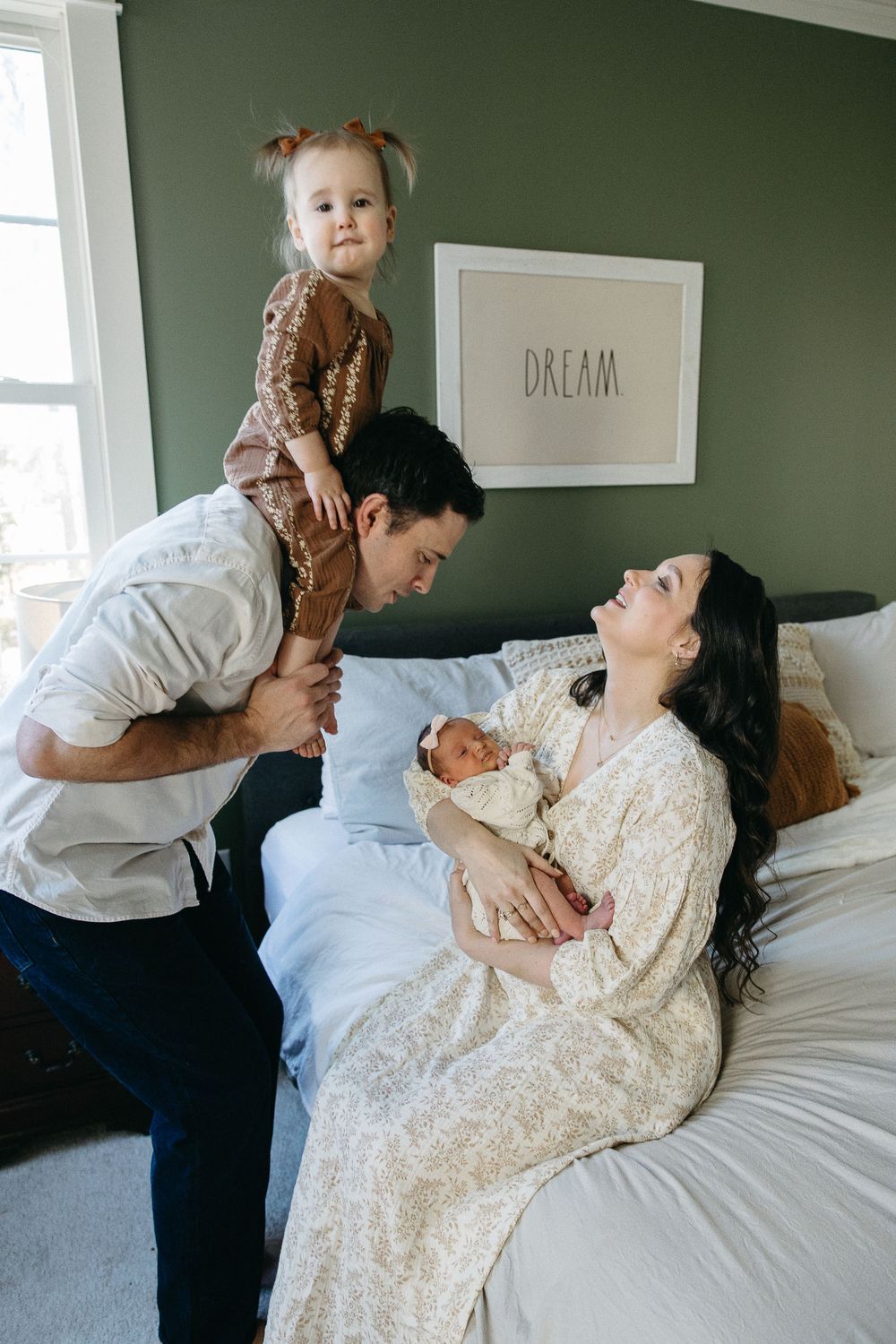 Family moment in bedroom with parents and children on bed next to DREAM sign on green wall.