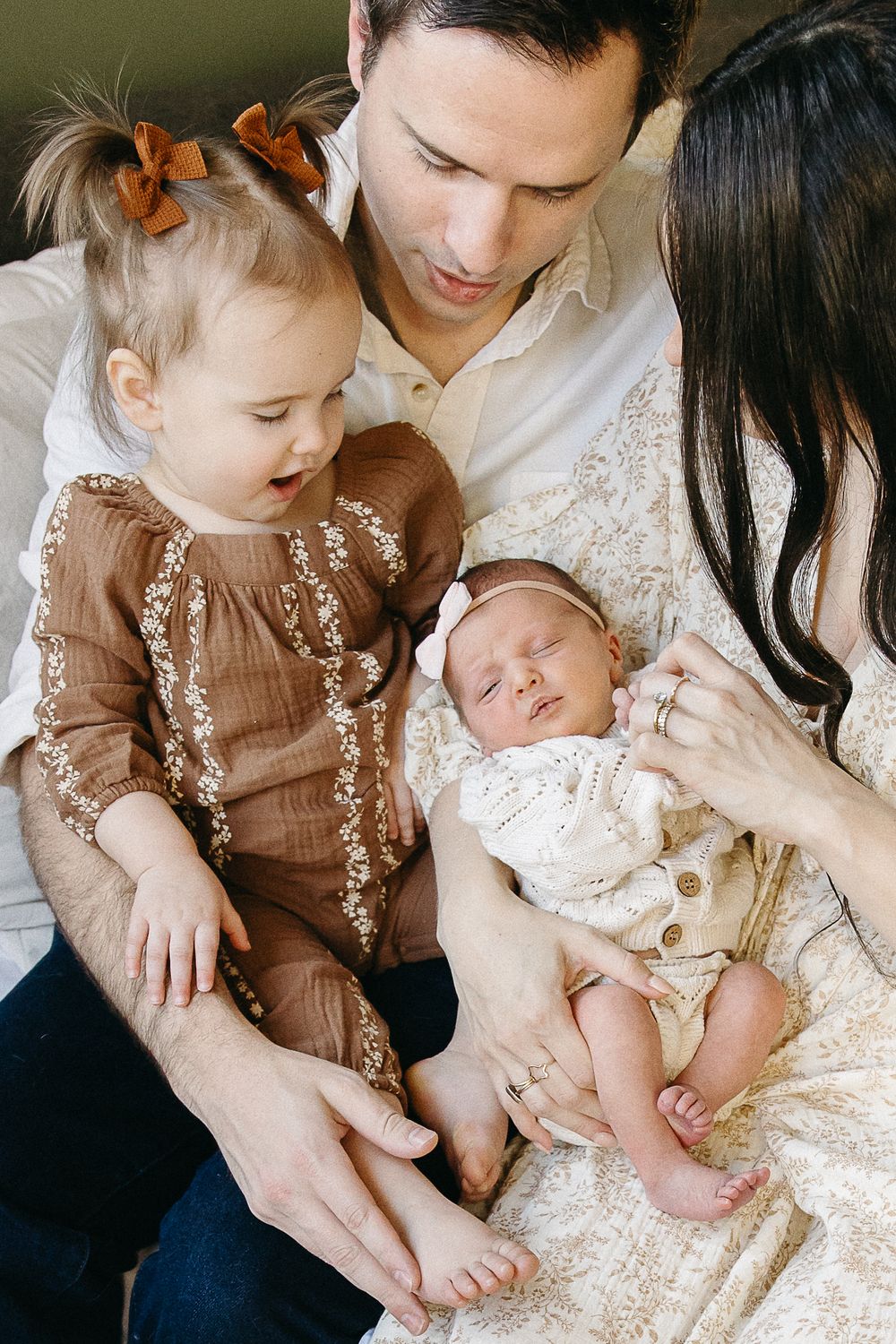 A family cuddles together on a couch, with a toddler in brown dress and newborn baby in white outfit.