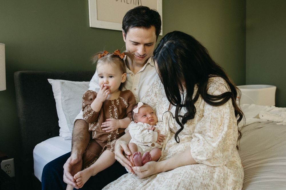Family sits together on dark couch in lace outfits with young child and newborn baby.