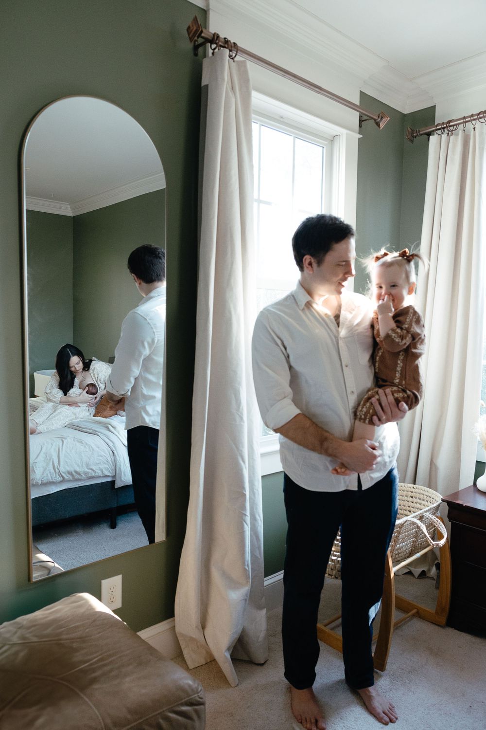 A nurturing moment captured in a bedroom with sage green walls, white curtains and a large mirror reflecting natural light.