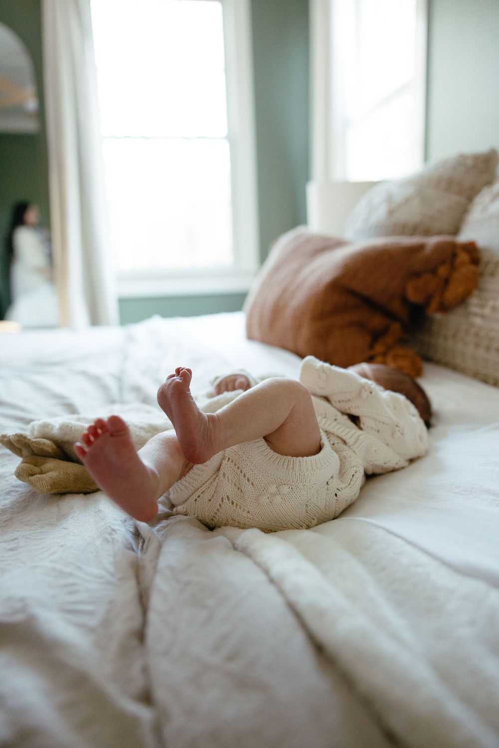 Small bare feet of baby wearing white knit dress laying on soft white bedding in sunlit room.