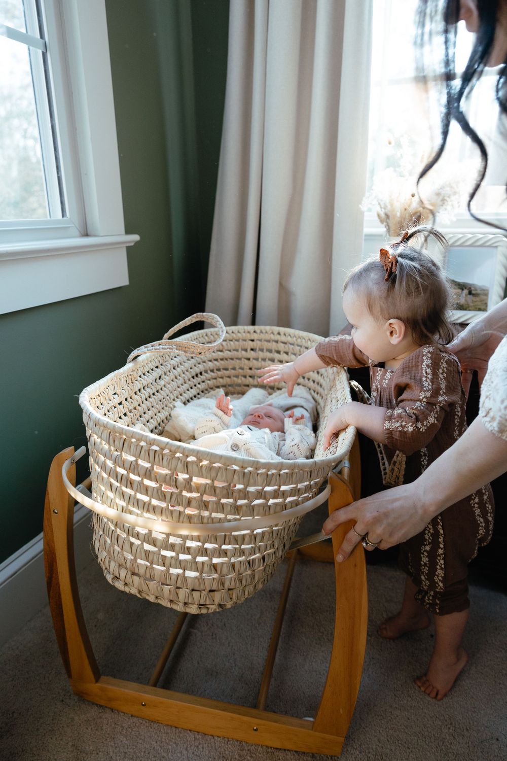 A modern nursery setting with a woven basket bassinet on a wooden stand beside a window with green walls.