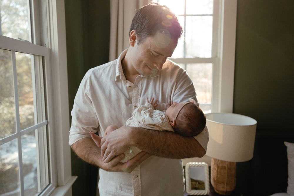 Someone in a white shirt cradles a newborn baby by a sunlit window with a table lamp nearby.