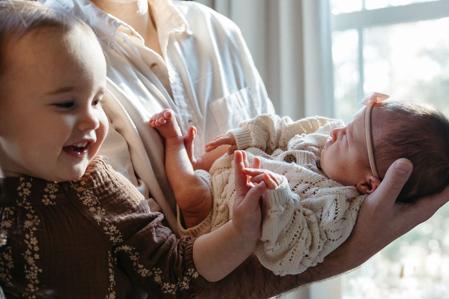 Toddler joyfully interacting with newborn baby sibling while being held in natural window light.