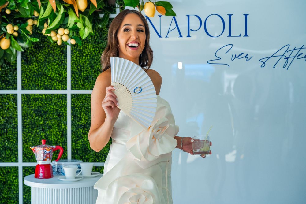 A festive celebration scene with white dress and decorative fan against a blue Napoli backdrop.