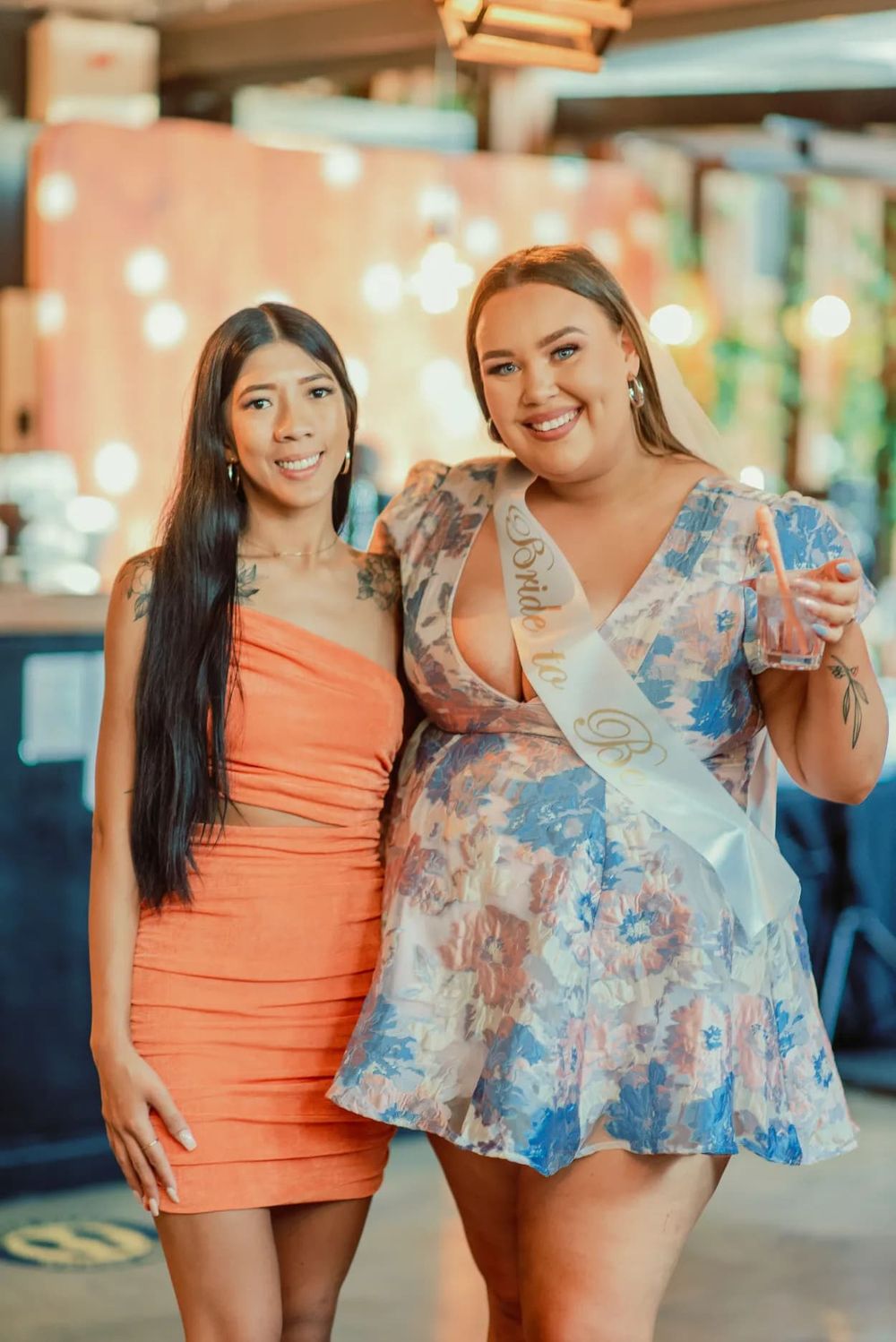 Two friends in colorful dresses pose together at an indoor event with bokeh lighting.