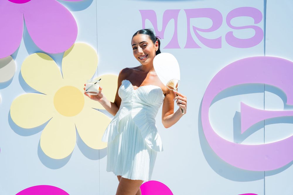 A person in a white sundress poses against a colorful wall with flower designs and balloons at an outdoor event.