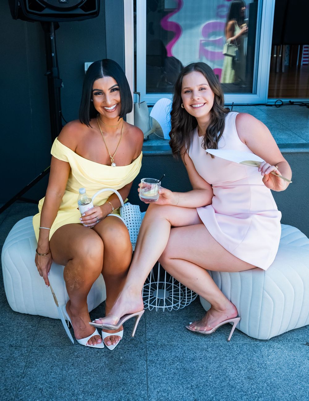 Two women in summer dresses sit on white outdoor furniture while holding beverages and chatting.