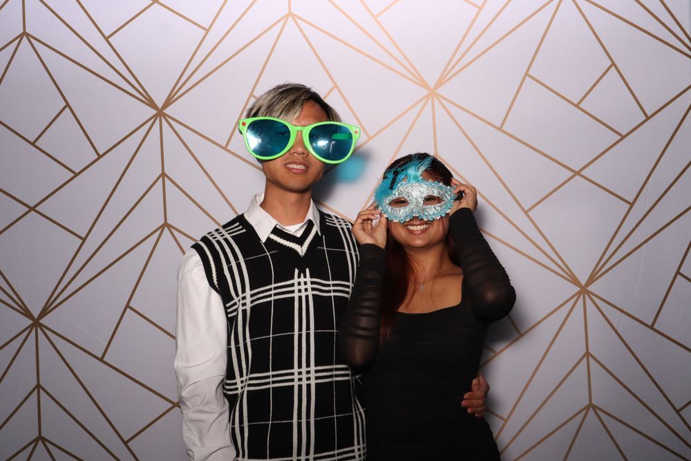 Two people wearing novelty sunglasses pose against a geometric gold and white backdrop.