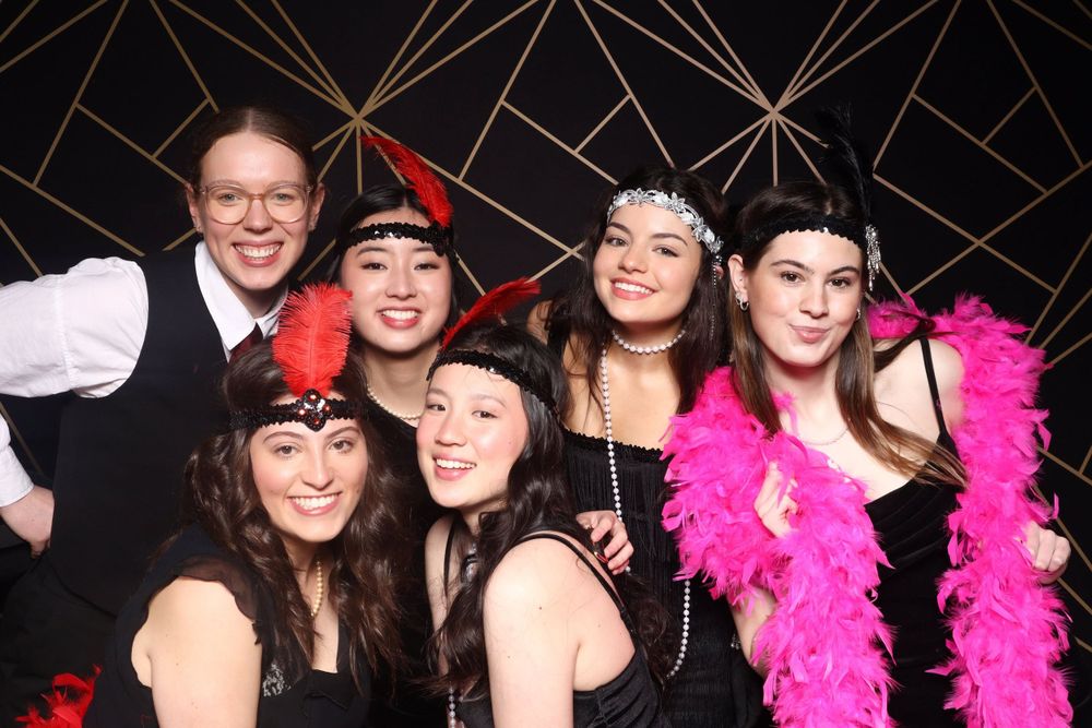 Photo booth party scene with group posing with feather boas and props against geometric backdrop.